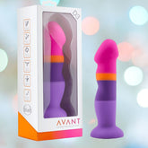 The Avant D3 7" Non-Porous Silicone Dildo - Summer Fling, featuring vibrant shades of pink, orange, and purple with a suction cup base, is showcased alongside its packaging. The box highlights the product's features and displays the brand name "Avant." A soft background with blurred lights emphasizes the body-safe design.